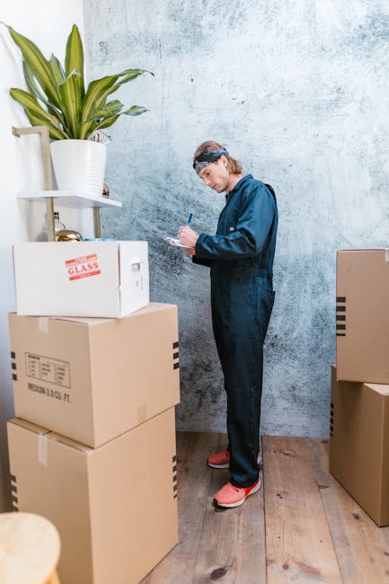 A person wearing a blue jumpsuit, protective eyewear, and pink footwear is inside a room, standing next to a stack of cardboard boxes. The individual is writing on a clipboard, likely documenting pack